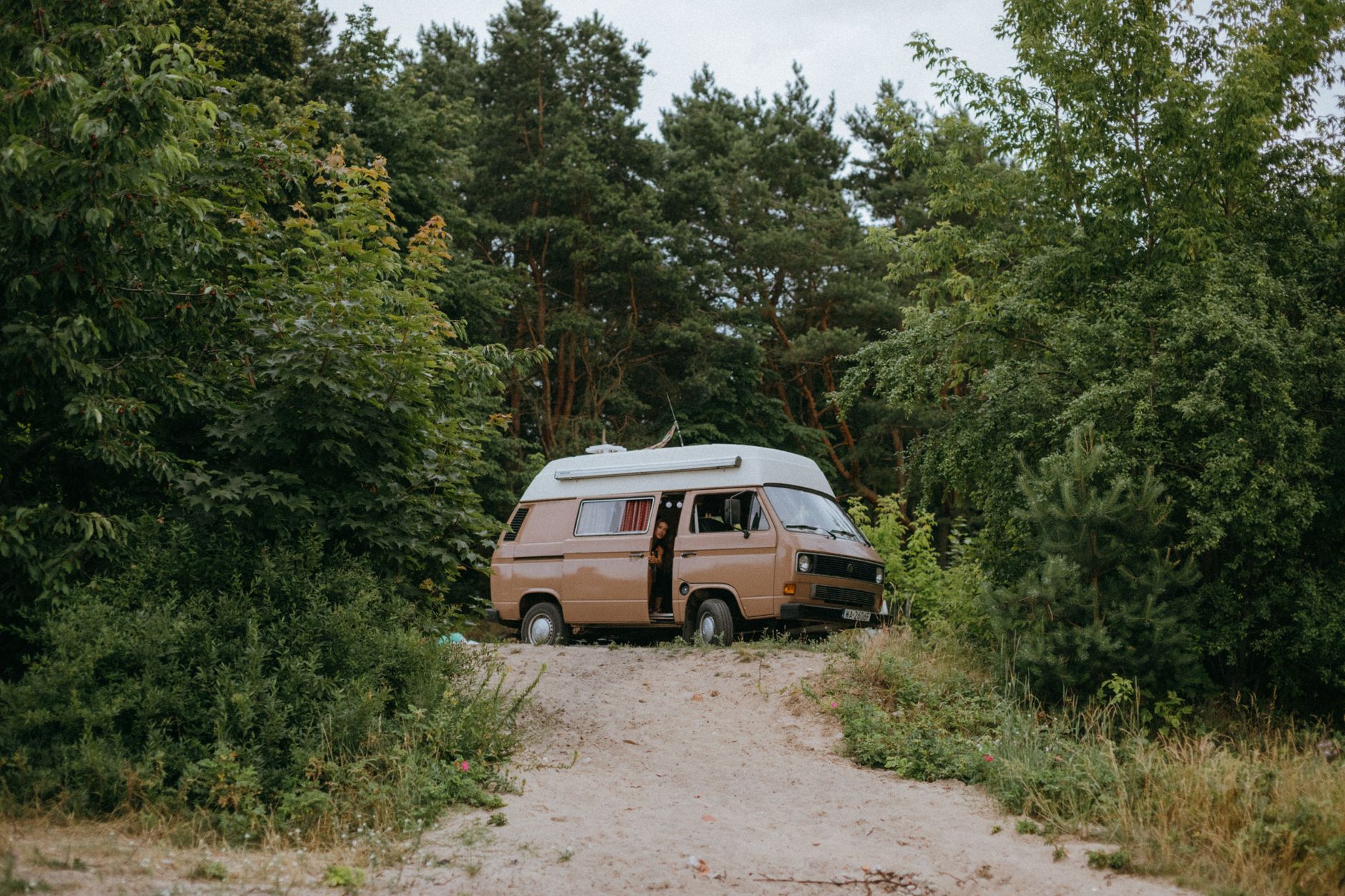 Camping-car beige à toit blanc garé sur un chemin de sable devant une forêt de pins, porte latérale ouverte.