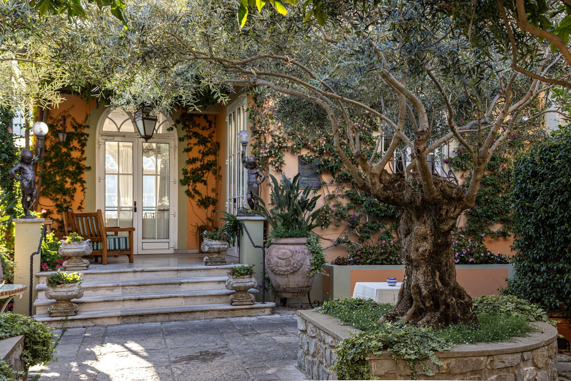 Cour d’une villa avec un vieil olivier, une façade ocre couverte de plantes grimpantes, un escalier de pierre, des vases et des statues en bronze.