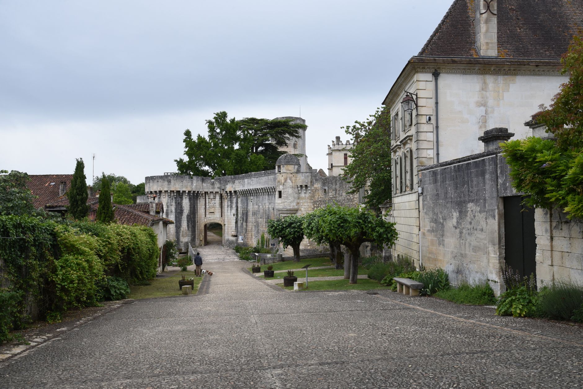 Rue pavée menant à une porte voûtée dans un rempart de pierre, entre des haies et une maison en pierre claire, sous un ciel gris.