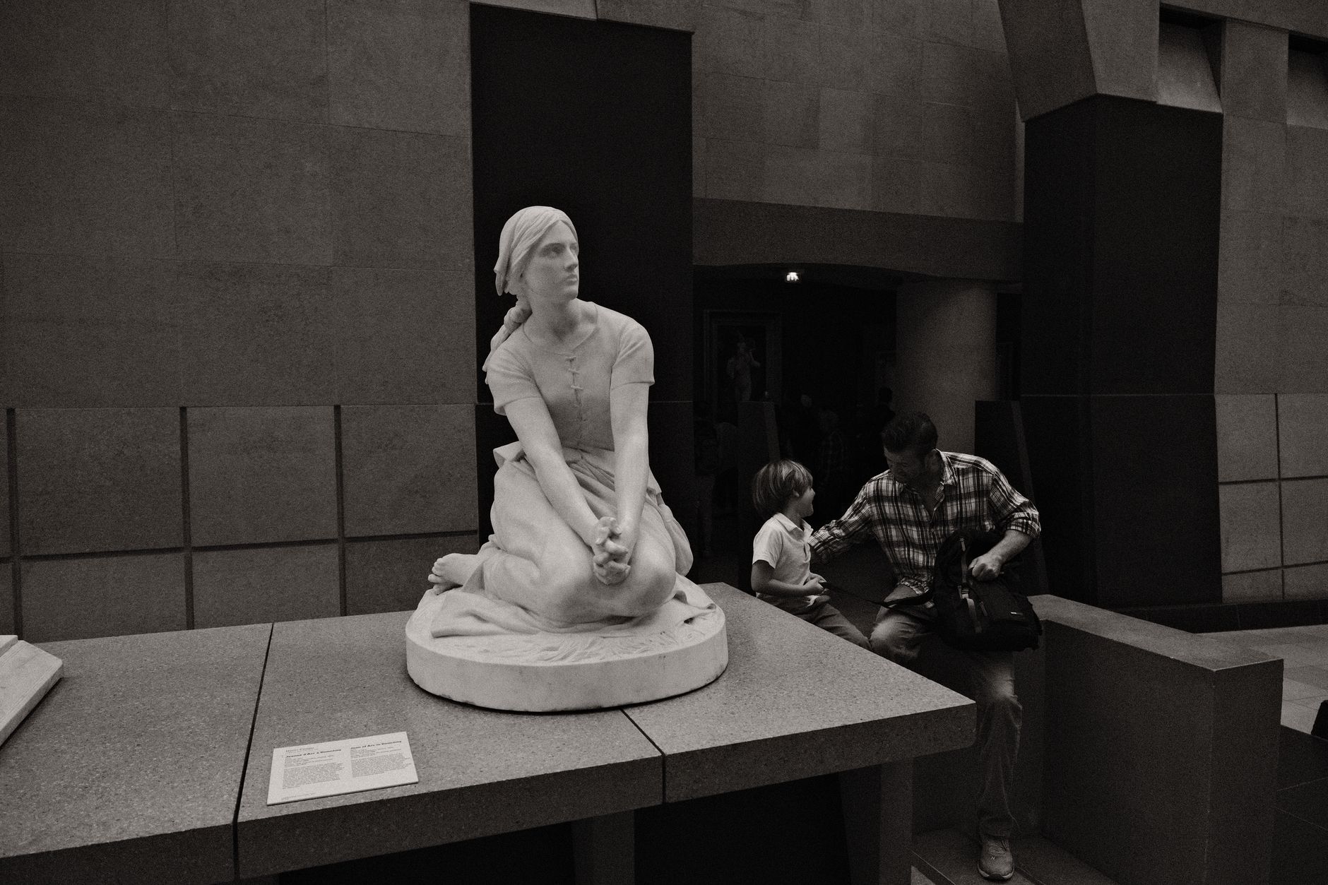 Statue d’une femme agenouillée dans une salle de musée, avec des visiteurs assis à proximité.