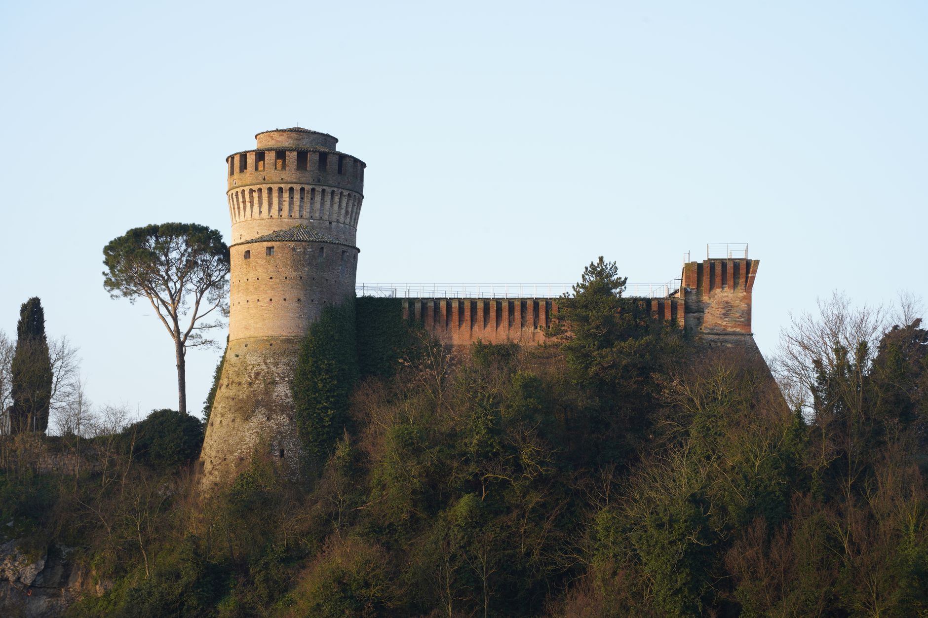 Tour ronde en brique et muraille crénelée d’un château au sommet d’une colline boisée, sous un ciel clair.